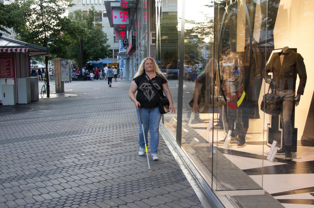 Grit, a blind woman, walks along the window of a shop with the White Cane IO.